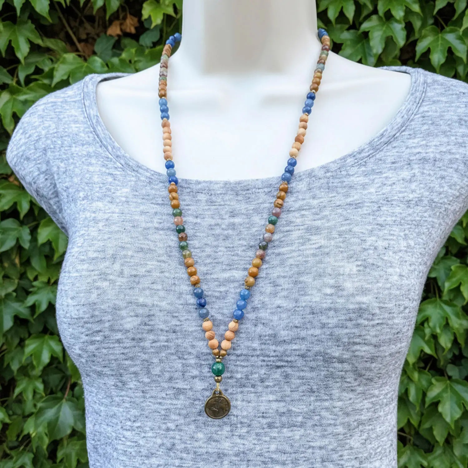 Mannequin wearing a long beaded necklace with a Earth pendant against a green leafy background