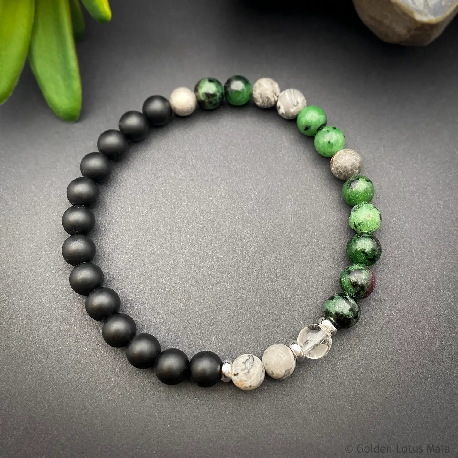 A beaded stretch bracelet arranged in a circle on a dark gray surface, featuring matte black Onyx, green Zoisite, marbled white and gray Jasper, and a clear faceted crystal bead. A small green leaf and stone are partially visible in the background.