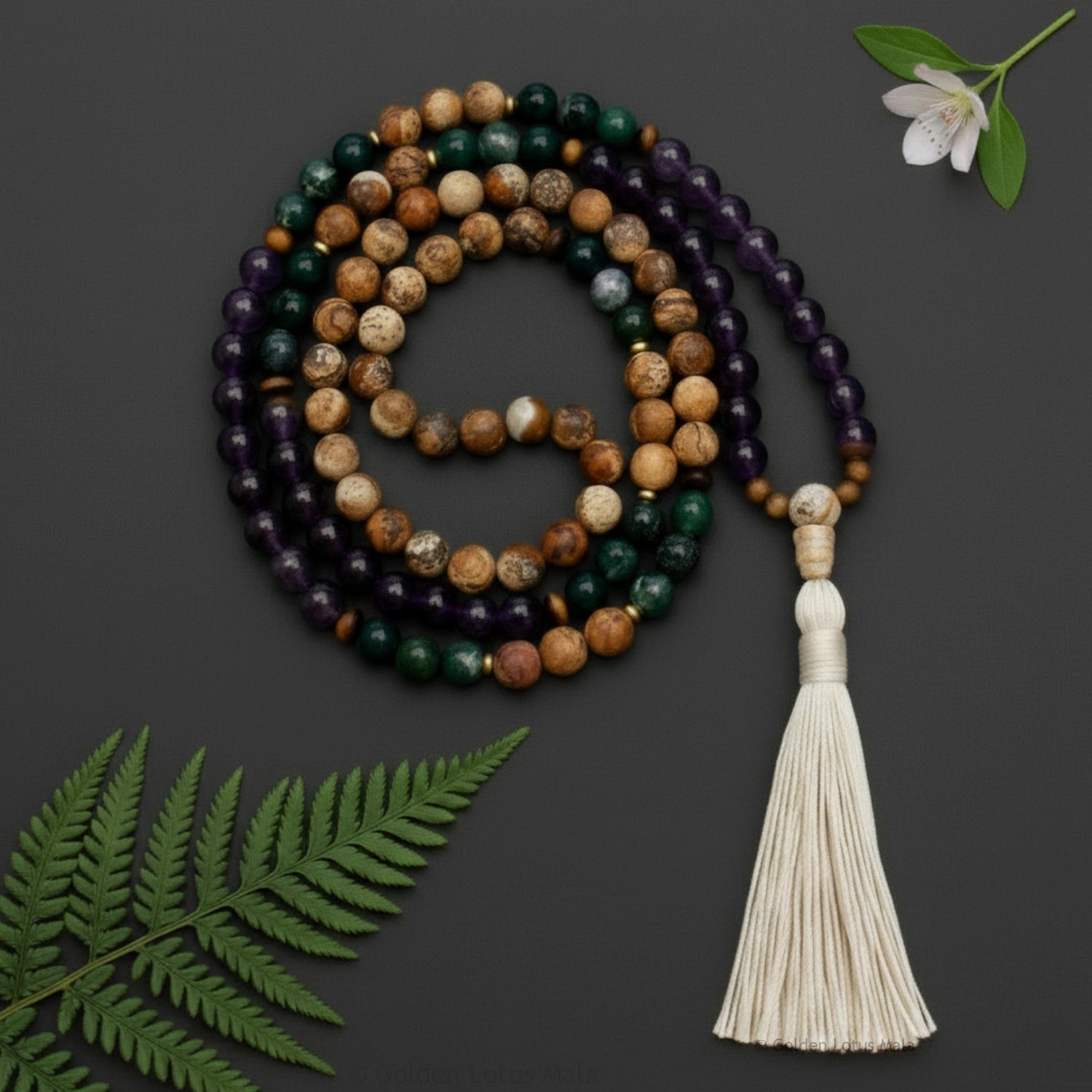 Gemstone mala necklace with tassel on a dark background with green leaves and a white flower.