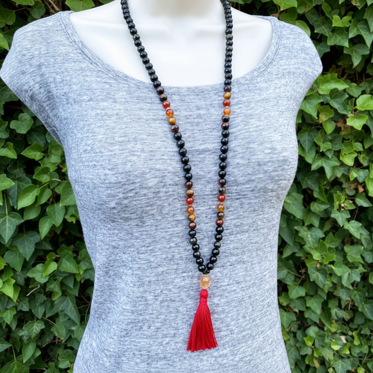 Mannequin wearing a long beaded necklace featuring a red tassel against a green leafy background.