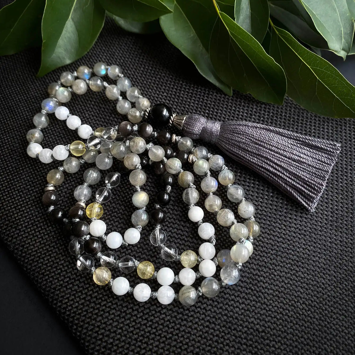 A grey and white mala necklace with dark gray tassel and accents of white and yellow.