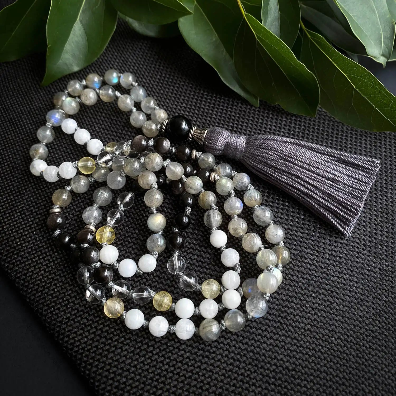 A grey and white mala necklace with dark gray tassel and accents of white and yellow.