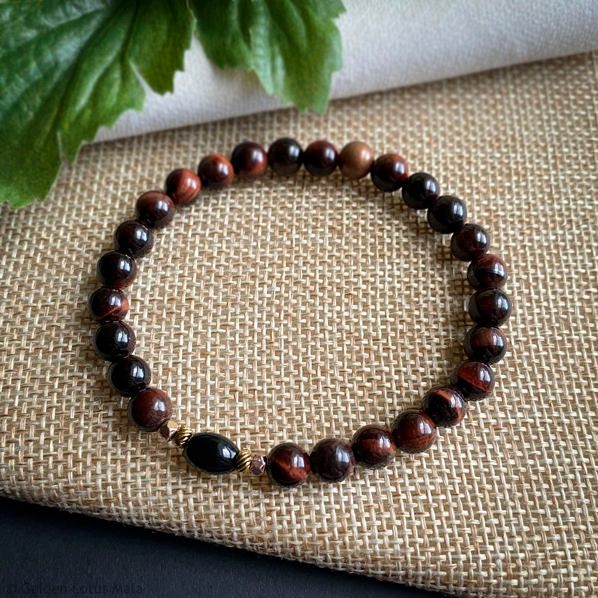 Reddish brown beaded bracelet on a textured fabric surface with green leaves in the corner.