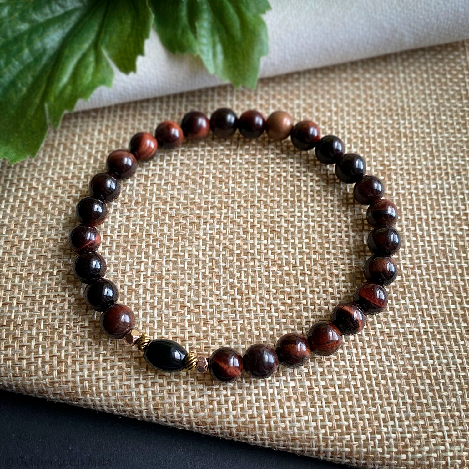 Reddish brown beaded bracelet on a textured fabric surface with green leaves in the corner.