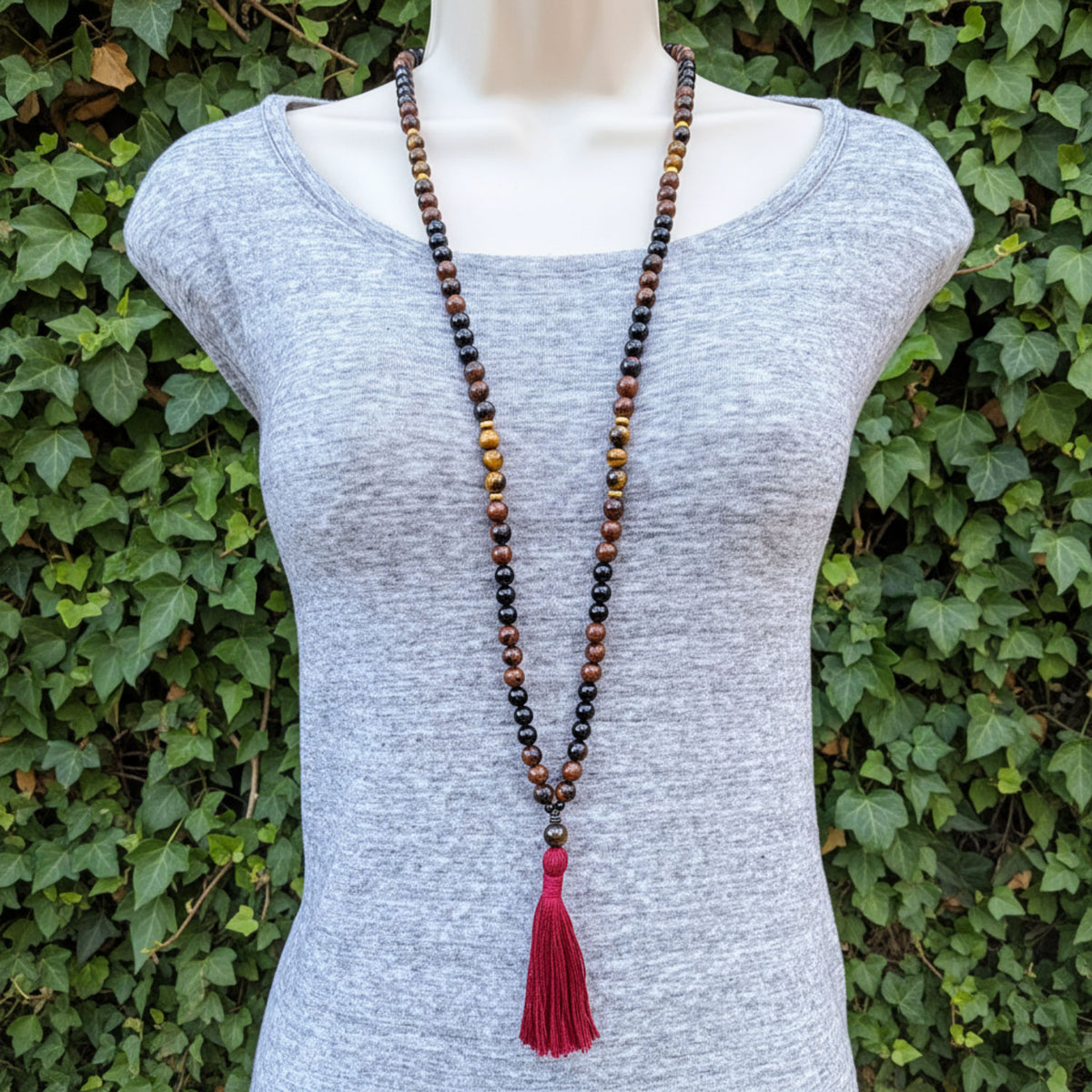 Mannequin wearing a gray shirt with a long beaded necklace featuring a pink tassel against a green leafy background.