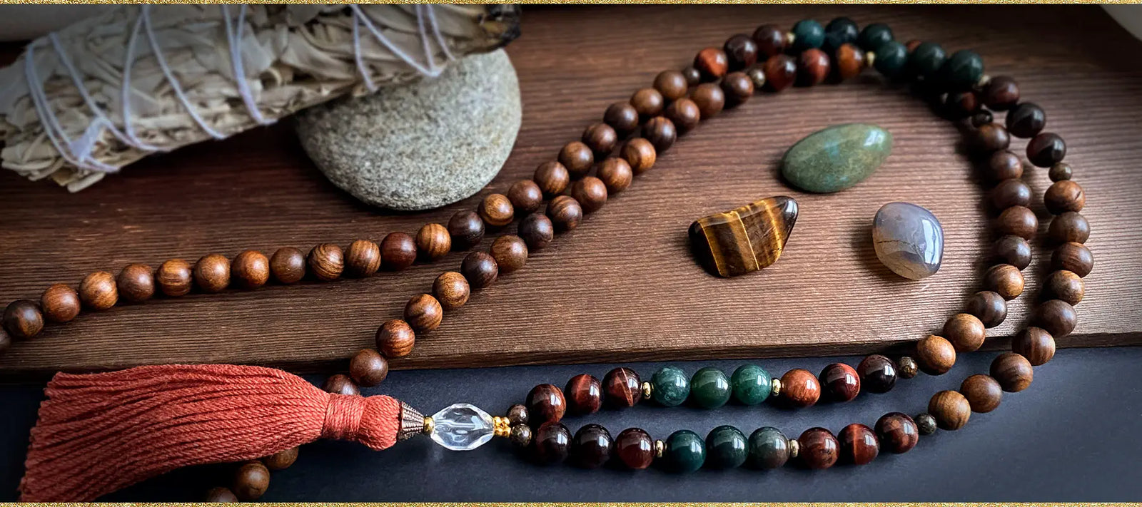 The Gemini Zodiac Mala necklace, made of Rosewood, Moss Agate, and Red Tiger Eye laying on a wood surface.